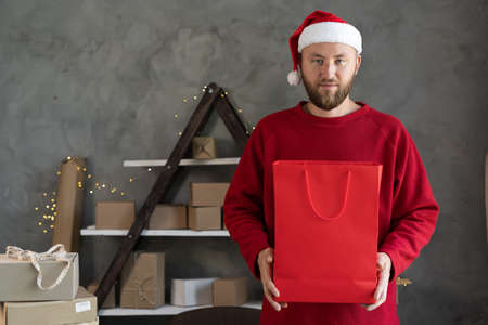 Caucasian bearded man in santa claus hat and red sweater gives red package with purchase or gift for New Year, delivery and logistics, online sellerの写真素材