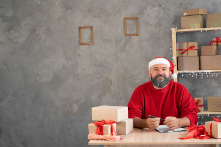 Smiling man, online store small business owner, manager, working entrepreneur in santa claus hat and red sweater sits at the table resting and lookingの写真素材