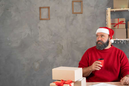 Millennial business owner online salesperson drinks coffee sitting at work, daydreaming and thinking Christmas. A man in a Santa hat with a glassの写真素材
