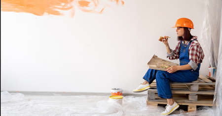 Woman repairman painted the wall with paint makes repairs and sits on a wooden pallet eating delicious pizza. Girl in an orange hard hatの写真素材