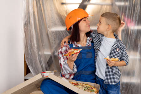 adorable family mom and son are resting and eating delicious Italian pizza tired after renovation in a new house. Caucasian woman in protective helmetの写真素材
