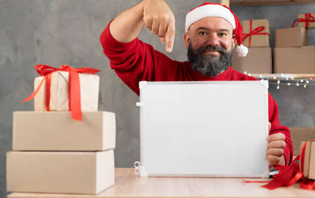 Santa in a red cap is working in the home office points his finger at a blackboard with a place for the text. Small business owner salesman Christmasの写真素材