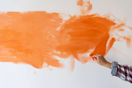 Renovation of the room, the white wall is painted with orange paint. Female hand with a paintbrush paints the wall at home indoors.repair concept.の写真素材