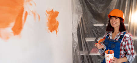 Happy woman in hard hat makes repairs and paints the wall in the new house with a brush in orange. Construction site and renovation concept.の写真素材