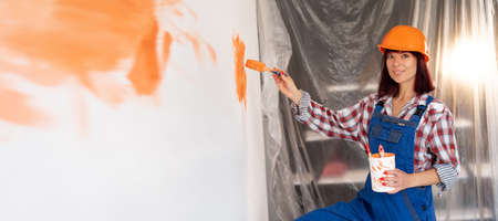 Happy caucasian woman in hard hat paints the interior wall in a new house with a brush in orange. Construction and renovation concept. Home renovationの写真素材