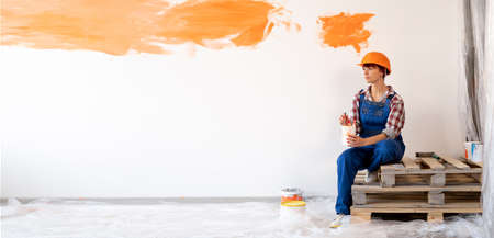 home renovation and wall painting. Caucasian woman with drawing brushes in a protective helmet and overalls sits resting on a wooden pallet.の写真素材