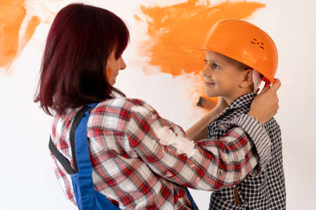 Repair. Happy family mom and son in helmet painting home interior wall. Paint the wall orange. Portrait of a woman with a child, repairman.の写真素材