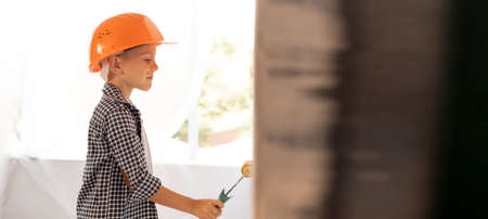 Caucasian boy in helmet paints a white wall with a roller in orange. A schoolboy helps to make repairs at home. spend time in quarantine.の写真素材