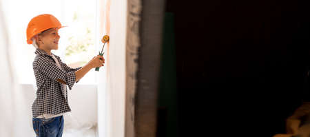 the child makes himself repairs in the room or at home. Boy in orange helmet painting the wall with a roller. Turnkey repair. Home renovationの写真素材