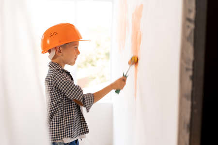 Caucasian child boy paints the wall with paint roller indoors doing repairs himself. children and home or apartment renovation. Kid in orange helmetの写真素材