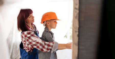 mom and son painting the wall together. repair apartments, a boy in an orange helmet and a woman whitewash the walls indoors. Home renovation conceptの写真素材