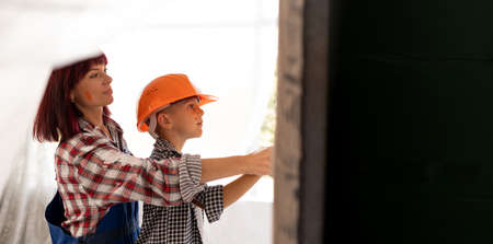 Mom teaches her son to do home repairs. Boy in orange helmet paint the wall in the room with a cushion helps the mother. A family makes renovationsの写真素材