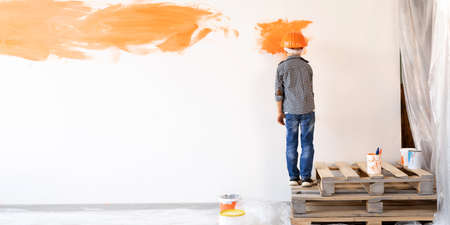 back view child paints a white wall in the room. House or apartment renovation. Children work in quarantine. A boy in an orange helmet with paintの写真素材