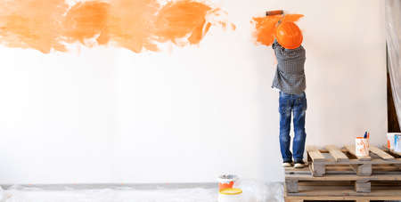 apartment renovation. Back view of a boy paints the wall with orange paint roller. The child helps to make repairs. Room renovation concept.bannerの写真素材