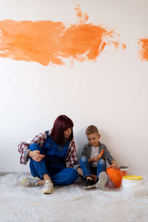 Renovation in the apartment. Happy family mother and son paint the wall with orange paint. Mom and boy sitting on the wooden floor are resting repairsの写真素材