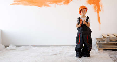 Turnkey repair. Little boy in a helmet and overalls with a paintbrush on the background of a white wall with orange paint. New home renovationの写真素材
