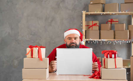 Small business owner bearded santa in a Christmas office or warehouse. Sad man in a Santa hat sitting at a table with a blackboard for text.の写真素材