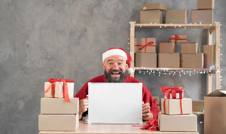 Happy bearded salesman in santa hat at home office. A man businessman holds a Christmas sale board in his hands. Internet delivery of goods.の写真素材