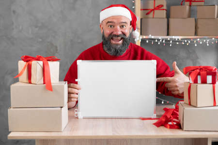 Christmas sale. The bearded small business owner points his finger at the place for the text. A man in a santa hat at the workplace warehouseの写真素材