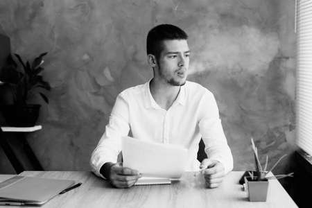young man in the office smokes a cigarette and exhales smoke while looking out the window thinking about the problem. business crisis and businessman.の写真素材