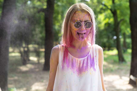 Blond woman at Holi festival of paints in a cloud of powder of multi-colored paint. The girl shows tongue to the crazy woman is having fun. Color Festの写真素材
