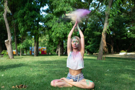 A blonde woman meditates in the park at the Holi festival raising her hands up sprinkling multi-colored dry paints. Outdoor meditation. colored powderの写真素材
