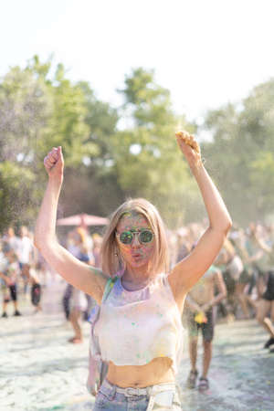Millennial girl dancing at the Color Fest festival of paints in the city park with her hands up. Traditional festival of dry urban paints. woman in sunglasses with dry paints faceの写真素材