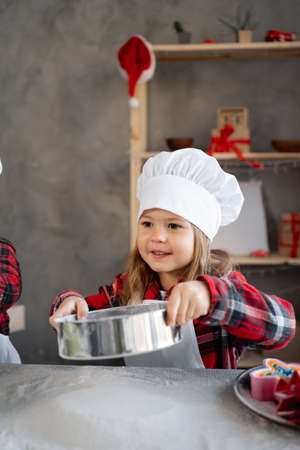 little girl sifts flour for dough through a sieve on the table. The child cooks in the chefs clothes. Children homemade cakes in the kitchen.の写真素材