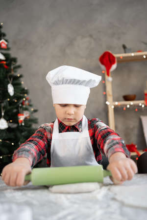 Serious boy in bakers uniform preparing cookies or pizza for Christmas. Child rolling out raw dough on the table. Homemade baking conceptの写真素材