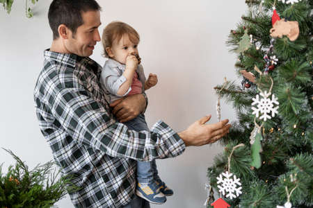 Caucasian dad and little son decorate the Christmas tree with toys. A man holds a child in his arms while standing near a fir tree on New Yearsの写真素材