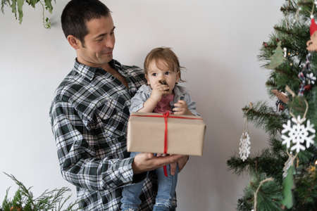 Young family dad and son celebrate Christmas at home near the fir. A man gives a box with a gift on Christmas eve to a little boy holding itの写真素材