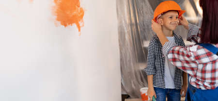new home renovation concept. Mom and son are painting the walls. Mother puts orange helmet on the boys head. Repair of apartments together. Banner.の写真素材