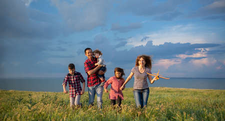 A large family with children launching a toy plane into the sky walking at sunset across a field with a blue sky background with clouds.の写真素材
