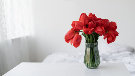 Glass vase with red tulip flowers in a light cozy bedroom interior. White wall, bed with white linen. Banner. Home vacation placeの写真素材