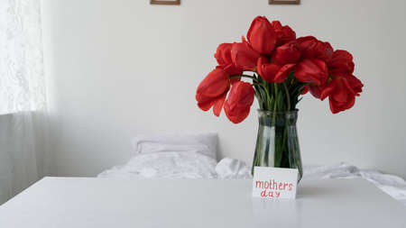 A bouquet of flowers in a vase for Mothers Day in the morning in the bedroom. Red tulips for Moms holiday and Happy Mothers Day greeting cardの写真素材