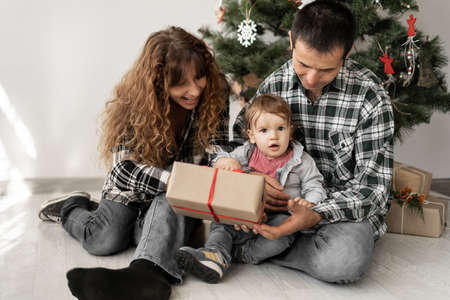 A young family with a baby on Christmas morning gives a gift in a box to their son. sitting on the floor under the tree. time together on New Years.の写真素材