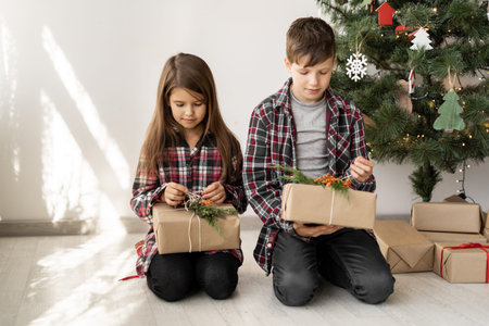 Children brother and sister with Christmas gifts are sitting on the floor of the room near the tree. Boy and girl look at the box with x-mas surpriseの写真素材