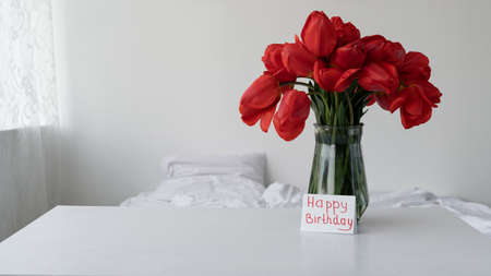 A bouquet of flowers in a vase for a birthday in the morning in the bedroom. Red tulips for the holiday and Happy birthday greeting card.の写真素材