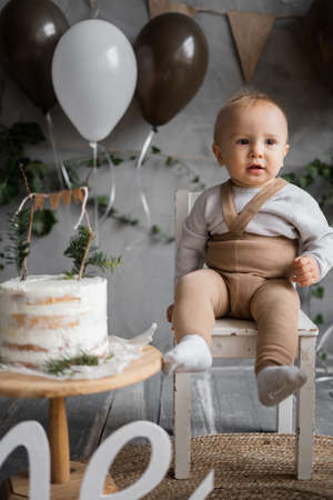 Babys first birthday, decorations with balloons and birthday cake, rustic minimalism style. Kid sitting on a high wooden chair, background.の写真素材