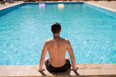 A man on vacation sunbathes in swimming trunks sitting by the pool with blue water, back view. Athlete with a beautiful body relaxes on vacation.の写真素材