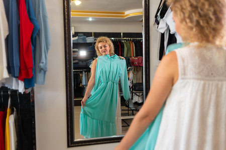 A blonde girl tries on a dress in front of a mirror in a clothing store, a fitting room in a womens clothing boutique, choosing and buying a dressの写真素材