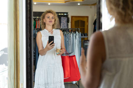 Woman customer buyer in a womens clothing store, photo on the phone in front of a mirror in a fashion boutique, sale in a mall.の写真素材