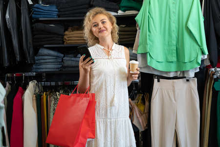 girl shopping in clothing store with paper bags, packed purchases in woman hands in fashion boutique, happy shopper on sale with coffee.の写真素材