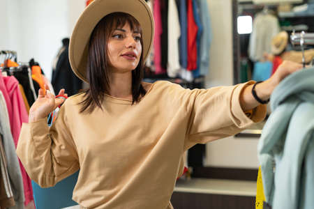 woman in a hat shopping in the clothing department of a mall, a girl choosing clothes on the hangers of a store, buying a product in a boutique.の写真素材