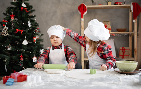 children prepare christmas cookies, boy rolls out dough, sister helps him by straightening hat, cooking gingerbread together, festive wall background.の写真素材