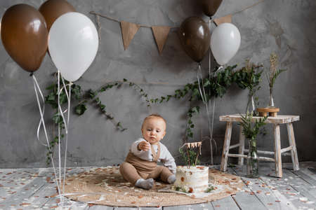 Festive decor for one year toddler, boy sitting on the floor with cake, gray background with brown and white balloons and natural materials decorations.の写真素材