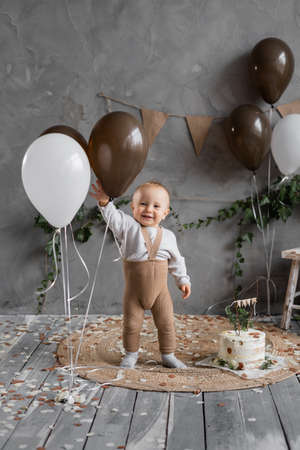 1st birthday, baby standing with balloons and a delicious cake on the floor, holiday decoration in rustic style, gray background, place for text.の写真素材