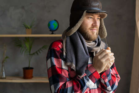 In winter, in a cold apartment without heating. A young Caucasian man wearing a hat and a scarf stands near the window and freezes. Close up portrait.の写真素材