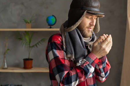 In winter, in a cold apartment without heating. A young Caucasian man wearing a hat and a scarf stands near the window and freezes. Close up portrait.の写真素材
