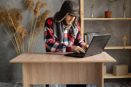 In winter, in a cold apartment without heating. A young Caucasian man wearing a hat and a scarf is working on a laptop at home in a cold apartment. Freelancer remotely online.の写真素材